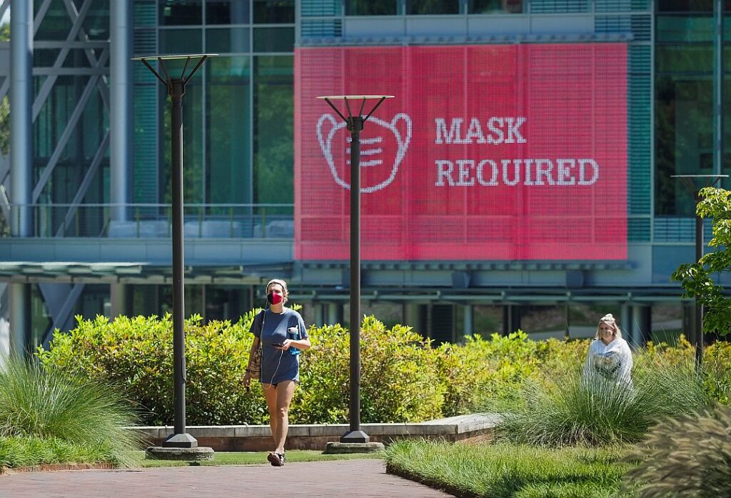 Masked student carrying phone with headphones walks in front of outdoor screen outside of Talley Student Union that has "mask required" projected. 